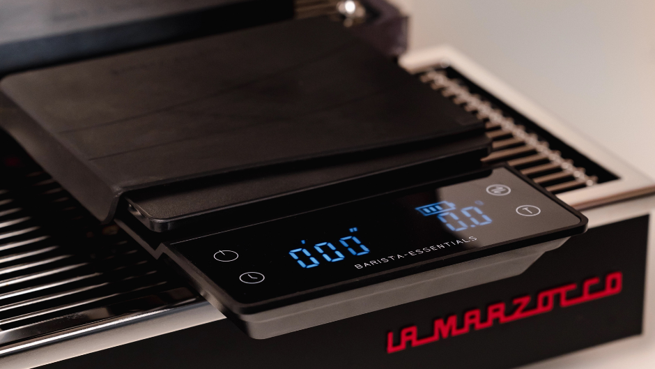 Zwarte koffieweegschaal met timer van Barista-Essentials, met display op 0,00 gram, op de afdruiprooster van een espressomachine.