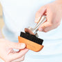 Image of a person holding a wooden brush and cleaning a stainless steel coffee tamper with it.