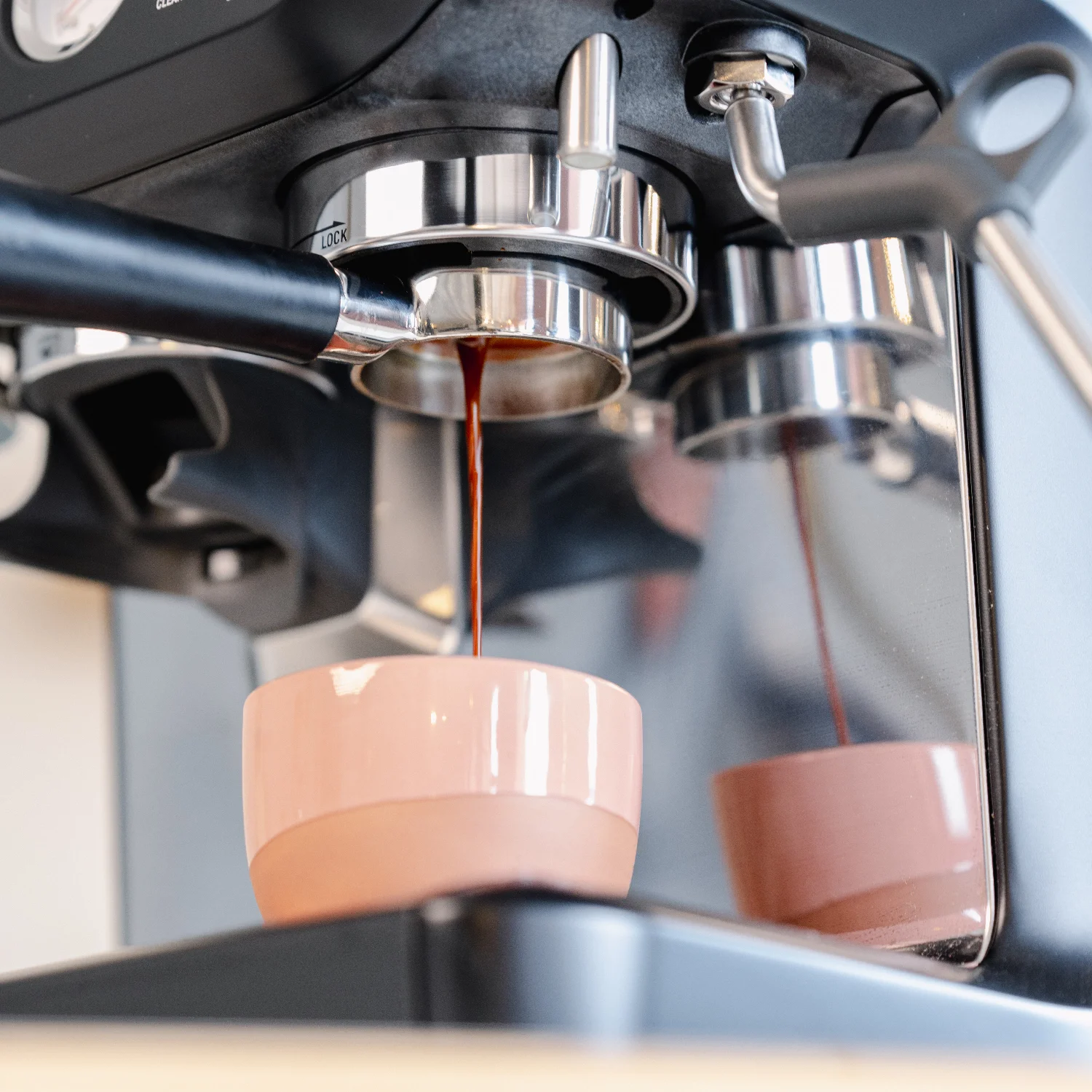 Een 54mm naked portafilter bevestigd aan een Sage espressomachine met een zwart handvat. Espresso stroomt in een roze kopje.