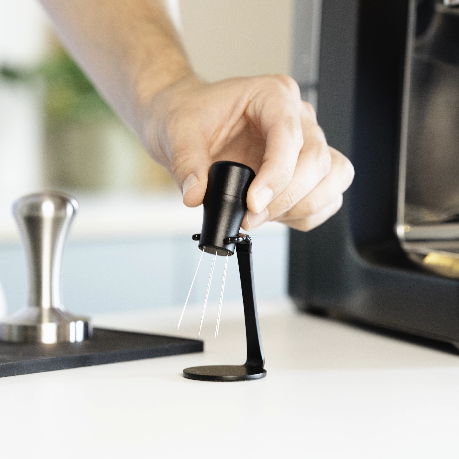 Een hand plaatst een espresso-distributietool met dunne metalen naalden in een zwarte standaard op een witte tafel, met een tamper en een espressomachine op de achtergrond.